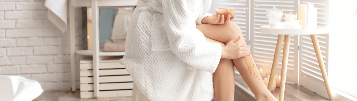 женщина в белом халате использует скраб для тела для ног в светлой ванной комнате